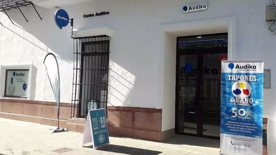 Fachada de un centro auditivo Audika con letreros promocionales. A la derecha, un cartel destaca “TAPONES de BAÑO a medida 50% DESCUENTO.’ Contexto: calle soleada frente a la clínica.