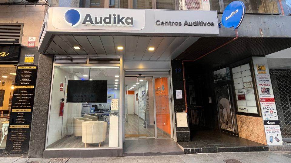 La fachada de un centro auditivo Audika muestra un salón iluminado con sillones beige y pantallas para atención al cliente. En el letrero superior se lee: 'Audika Centros Auditivos'.