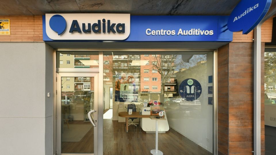 La fachada de una clínica auditiva Audika muestra un letrero azul con el texto ’Audika Centros Auditivos.’ A través de la cristalera, se aprecia un interior moderno con mesas, sillas y material promocional de audífonos. En el entorno urbano, se reflejan edificios residenciales. Texto visible: ’Audika,’ ’Centros Auditivos,’ ’59€.’