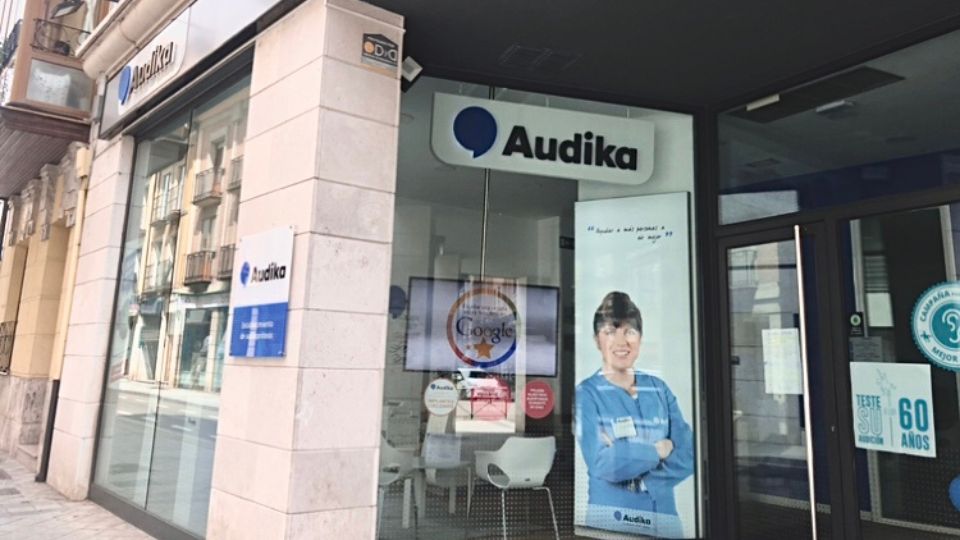 Fachada de una clínica Audika especializada en el cuidado auditivo, con ventanales que exhiben publicidad relacionada y pegatinas de certificaciones. Entorno urbano con edificios residenciales al fondo.