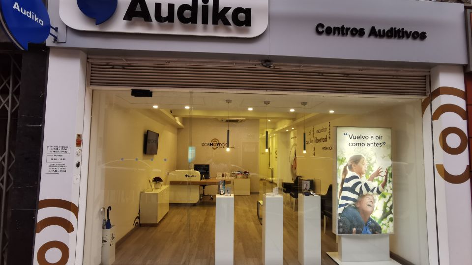 Fachada de un centro auditivo Audika con diseño moderno, mostrador interior visible, vitrinas con productos y un cartel que dice: 'Vuelvo a oír como antes'.