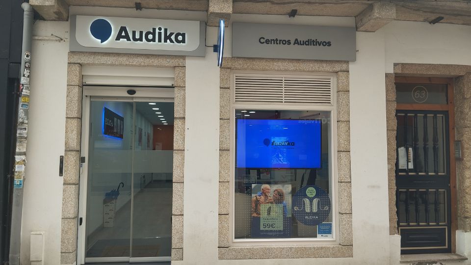 La fachada de un centro auditivo Audika muestra: puerta de entrada de vidrio, ventana con cartel promocional de audífonos desde 59€, interior iluminado y letrero ’Centros Auditivos’.