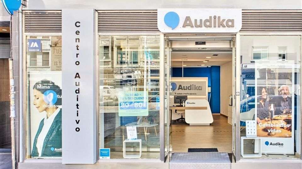 Fachada de un centro auditivo Audika con diseño moderno. A la izquierda, un cartel dice ’Mima tus oídos’. En el centro, recepción minimalista; a la derecha, letrero: ’Cambiamos vidas’.