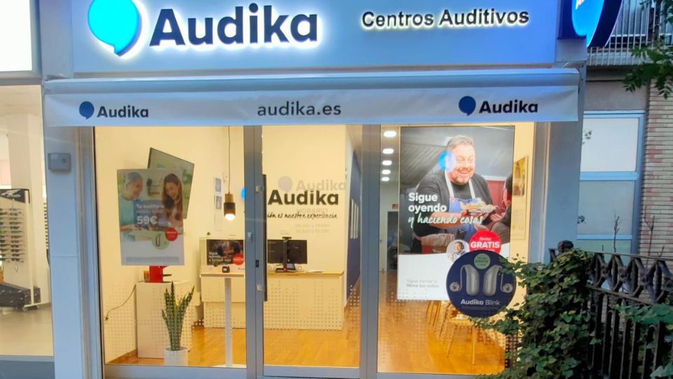 Fachada de un centro auditivo Audika iluminada, con logotipo azul y cartelera promocional. Textos visibles incluyen: “audika.es’, ’Sigue oyendo y haciendo cosas’, ’Audífonos desde 59€’, ’Audika Blink GRATIS’. Interior ordenado.