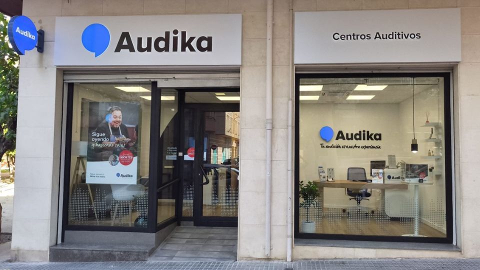 Fachada de un centro auditivo Audika con dos vitrinas. La izquierda muestra un cartel que dice: ’Sigue oyendo. Precisamos cuidarte. Gratis: prueba auditiva.’ La derecha revela un interior moderno con escritorio, silla y materiales promocionales, bajo el lema: ’Tu audición, nuestra experiencia.’