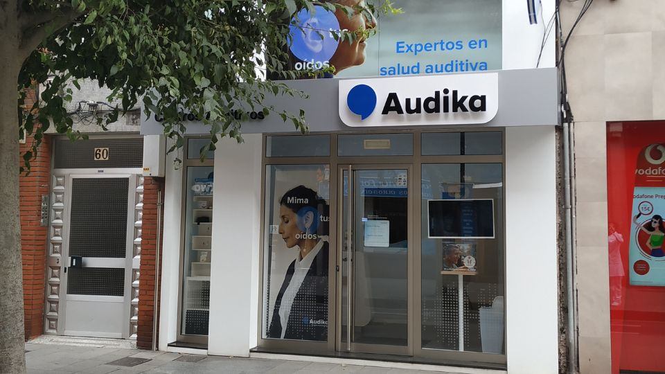 Fachada de una clínica auditiva Audika con gran cartel que dice “Expertos en salud auditiva.’ Contexto urbano, incluye puerta numerada 60 a la izquierda y comercio Vodafone a la derecha.