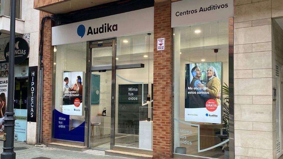 Fachada de una clínica auditiva Audika con cristales transparentes que muestran carteles promocionales: ’Mima tus oídos’ y ’Me encantan estos sonidos. Revisión GRATIS’. Entorno urbano con negocios cercanos.