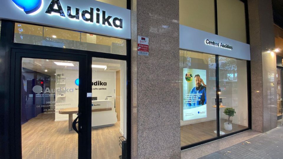 Fachada de un centro auditivo Audika con letreros luminosos. En el escaparate, un póster promocional y plantas decorativas. Horario en la puerta: **09:30h-13:30h, 16:00h-19:00h** (lunes a viernes).
