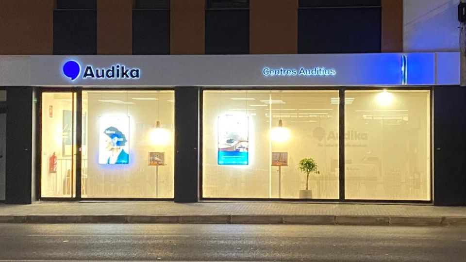 Fachada nocturna de una clínica auditiva Audika con letrero iluminado “Audika Centres Auditius.’ Ventanales muestran carteles promocionales y decoración modernas.