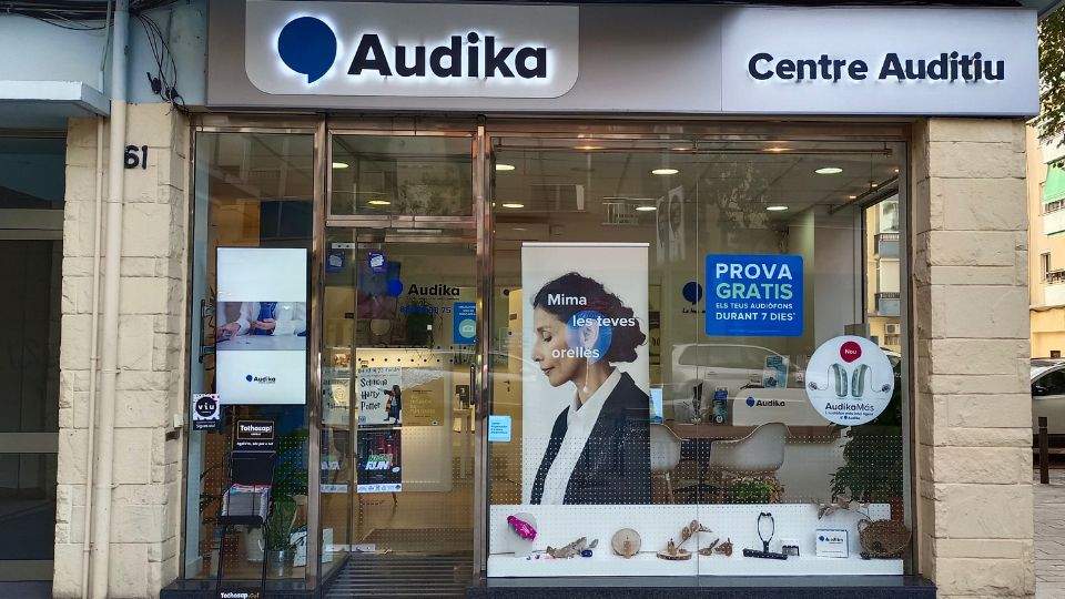 Fachada de una clínica auditiva Audika con el rótulo ’Centre Auditiu’. En el escaparate destacan carteles como ’PROVA GRATIS ELS TEUS AUDÍFONS DURANT 7 DIES’ y productos relacionados con audífonos.