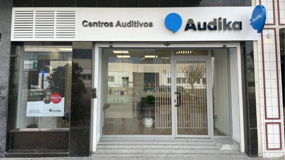 Fachada de un centro auditivo Audika con letrero que dice ’Centros Auditivos.’ A la izquierda, un cartel promociona audífonos con texto: ’Sigue oyendo y riendo. GRATIS prueba de audición.’