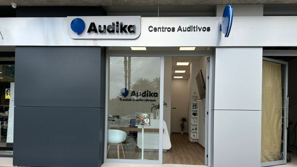 La entrada de un centro auditivo Audika muestra un letrero que dice ’Audika Centros Auditivos’. En el interior, hay muebles blancos, suelo de madera y equipo audiológico visible. Texto adicional en la ventana: ’Tu audición, es nuestra experiencia.’ El contexto incluye reflejos de palmeras y un entorno urbano.