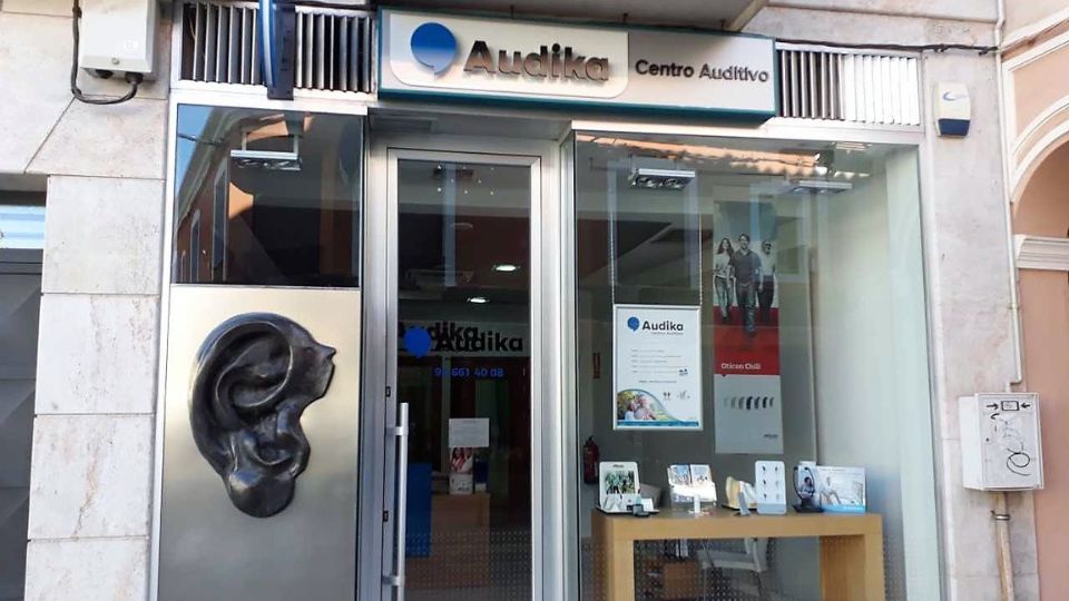 La fachada de un **centro auditivo Audika** muestra una escultura en relieve de una oreja a la izquierda. En el escaparate destacan folletos, vitrinas con audífonos y un póster que menciona **’Audika CROS’**. El contexto es una calle comercial.