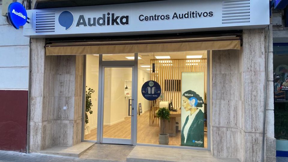 Fachada de una clínica auditiva Audika con cartel ’Centros Auditivos’ en la parte superior. Ventanas muestran interiores iluminados con decoración moderna y textos: ’Audika | Estás en buenas manos’ y ’Mima tus oídos’.