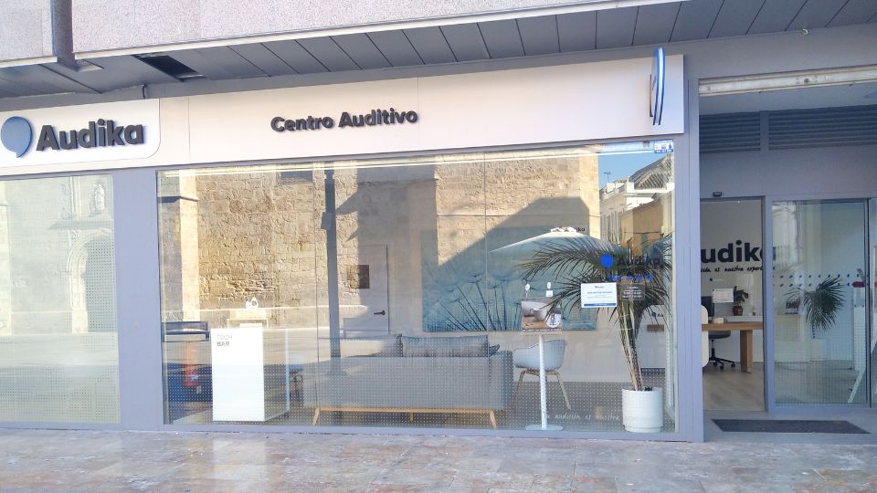 Fachada de un centro auditivo Audika con letrero ’Centro Auditivo’, grandes ventanales mostrando interior moderno con muebles minimalistas y planta decorativa. Textos visibles: ’TOUCH B&F’ y ’audición a medida’. Ubicación iluminada en calle urbana.
