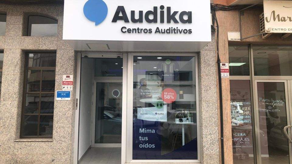 Entrada de un centro auditivo Audika con letrero que dice “Audika Centros Auditivos.’ En el escaparate se leen mensajes: “Tu edad igual a tu descuento,’ “58 años - 58%,’ y “Mima tus oídos.’ Su entorno incluye edificios contiguos con carteles de negocios vecinos.