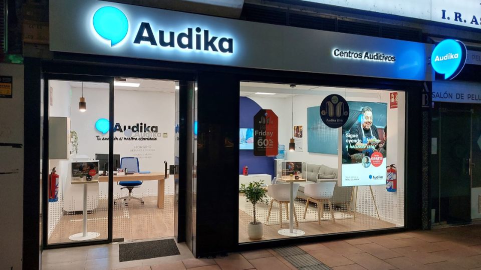 Fachada iluminada de un centro Audika con el letrero ’Centros Auditivos’. En el interior, se observa una sala de espera moderna y un escritorio. Texto destacado: ’Sigue oyendo y hazlo gratis’ (Audika) y ’Friday 60%’.