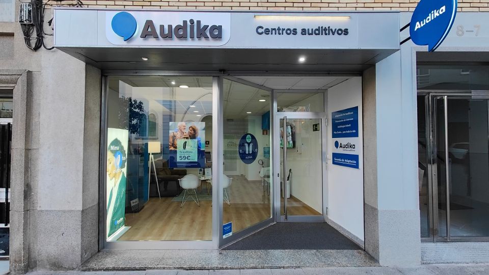 Fachada de un centro auditivo Audika con cristaleras que permiten ver un interior moderno. Carteles promocionales destacan: ’Audífonos desde 59€’ y ’Mimamos tus oídos.’