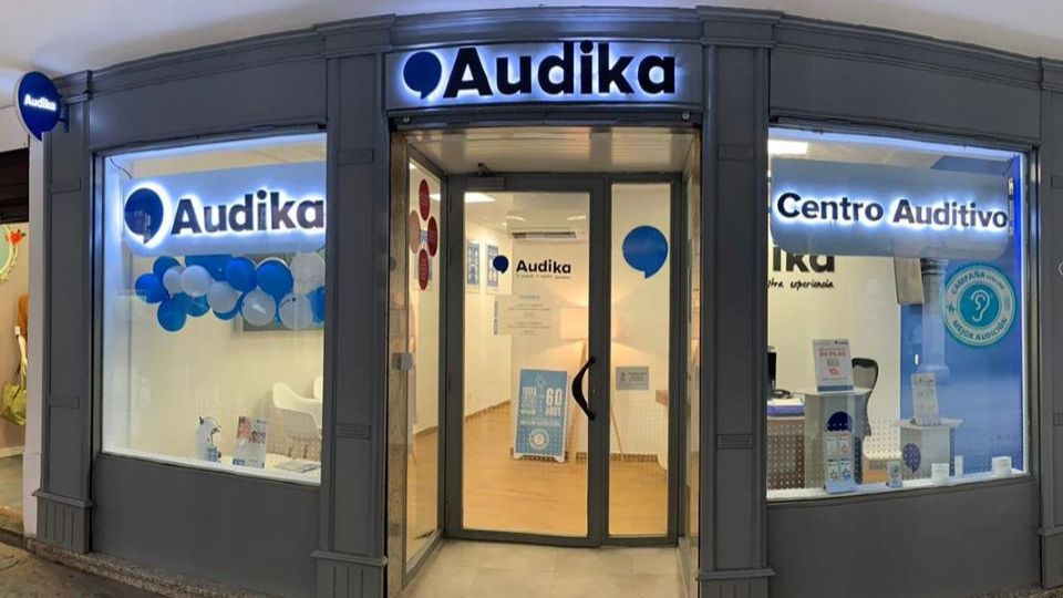 Fachada de una clínica auditiva Audika con letreros luminosos “Audika’ y “Centro Auditivo’. Interior moderno con decoración blanca y azul; ofrece pruebas de audición y servicios para audífonos.