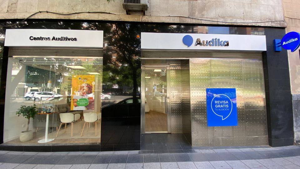 Fachada de un centro auditivo Audika con letreros que incluyen: ’Centros Auditivos’ y ’REVISA GRATIS TU AUDICIÓN.’ Al interior, se ven sillas blancas y póster de Audika.