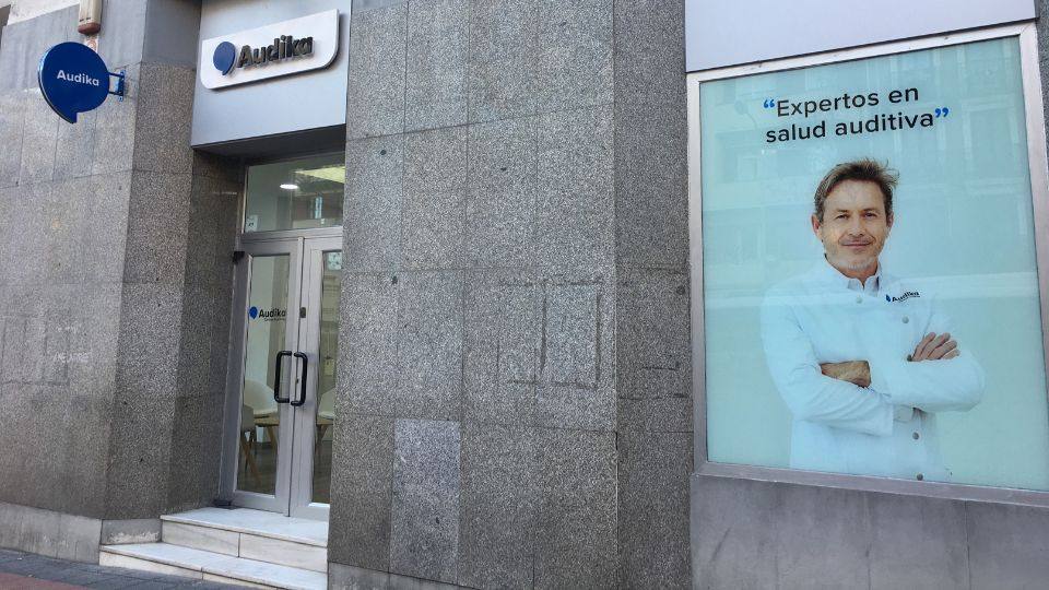 Fachada de una clínica auditiva Audika con entrada de vidrio, logotipo visible y cartel promocional mostrando un profesional con bata, acompañado del texto: “Expertos en salud auditiva’.