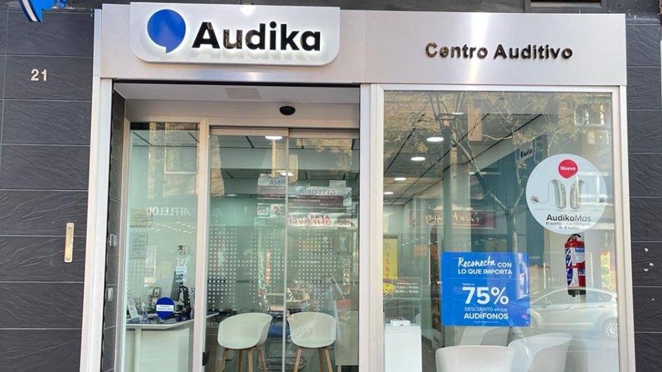 Entrada de un centro auditivo Audika con fachadas de vidrio, logotipo visible y texto: ’Audika. Centro Auditivo.’ Promoción anunciada: ’75% descuento en tus audífonos. Reconéctate con lo que importa.’ Panel lateral destaca ’AudikaMás,’ un servicio nuevo. Interior moderno y luminoso.