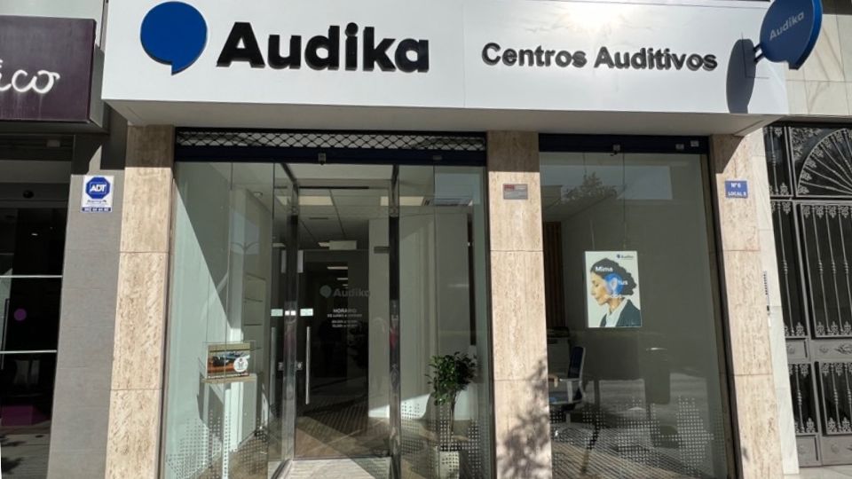 Fachada de una clínica Audika Centros Auditivos con letrero visible. Acceso con puertas de vidrio, interior iluminado con mobiliario moderno. A la derecha, un cartel promociona servicios de audición.