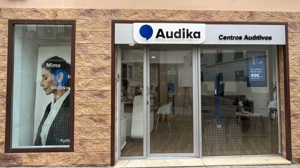 Fachada de un centro auditivo Audika con ventanas de cristal y un cartel promocional que dice: ’Tu audífono desde solo 49€’. Interior moderno con muebles claros.