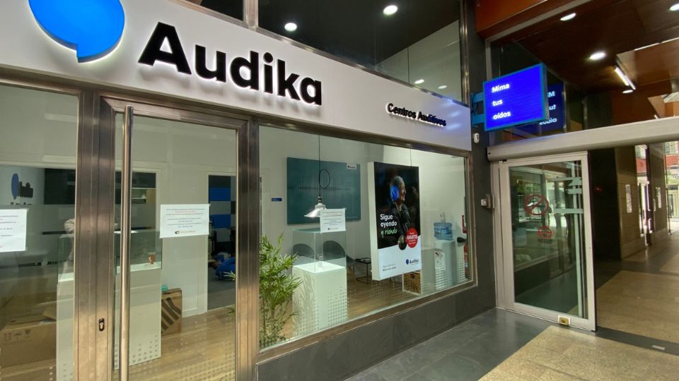 La fachada de un centro auditivo Audika muestra cristales amplios que revelan un interior moderno con muebles minimalistas. El cartel dice: “Sigue oyendo el ritual’ junto a un logo de Audika. En la esquina superior, un letrero digital azul muestra “Mima tus oídos.’