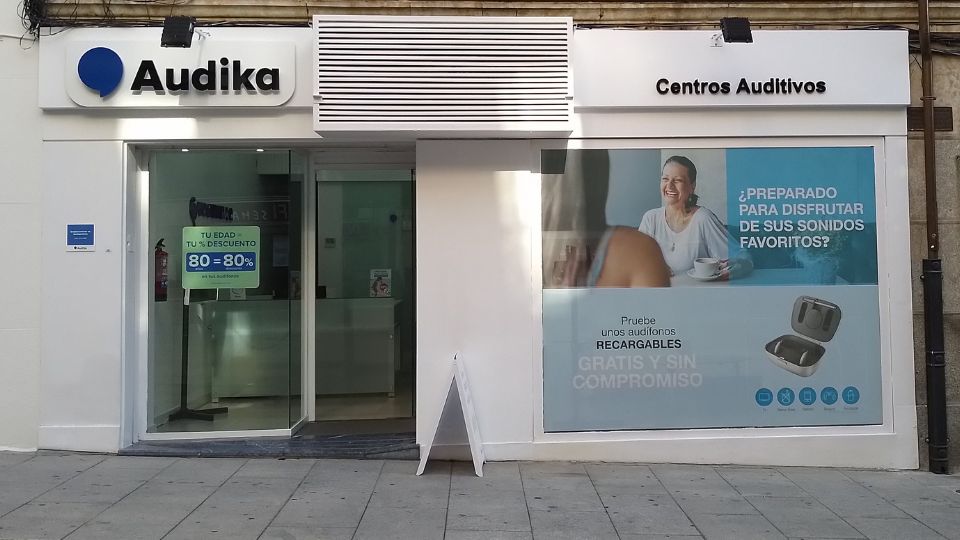 Fachada de una clínica auditiva Audika con letrero ’Centros Auditivos’. En el escaparate, oferta: ’Tu edad, tu descuento 80=80%’. Cartel publicitario: ’¿Preparado para disfrutar de sus sonidos favoritos? Pruebe unos audífonos recargables GRATIS Y SIN COMPROMISO’. Visible imagen de audífonos recargables y logotipos asociados. Contexto: calle peatonal.