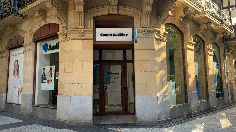 Fachada de un centro auditivo Audika con letrero ’Centro Auditivo’ sobre la puerta principal. Ventanas muestran publicidad: ’Tu vida a otro nivel’ y servicios de audífonos. Contexto urbano.