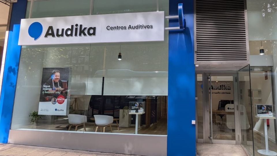 Fachada de una clínica auditiva de Audika con señalización ’Centros Auditivos’. En el interior hay sillas blancas y un cartel promocional que dice: ’Sigue oyendo tu mundo con GRATIS* Prueba auditiva’. Ambiente urbano con árboles reflejados en el cristal.
