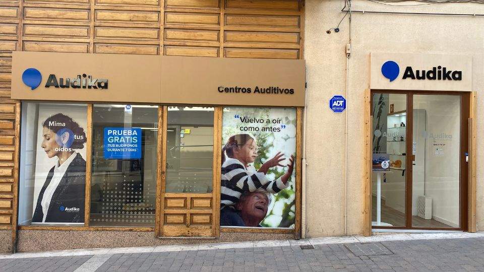 Fachada de un centro auditivo de Audika con letreros que incluyen: ’Mima tus oídos’, ’PRUEBA GRATIS tus audífonos durante 7 días’ y ’Vuelvo a oír como antes’. Ambiente urbano.