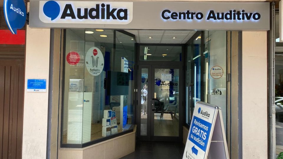Fachada de un Centro Auditivo Audika, destacado con un cartel ofreciendo ’Revisamos GRATIS su audición’. El escaparate muestra audífonos y servicios relacionados con la salud auditiva.