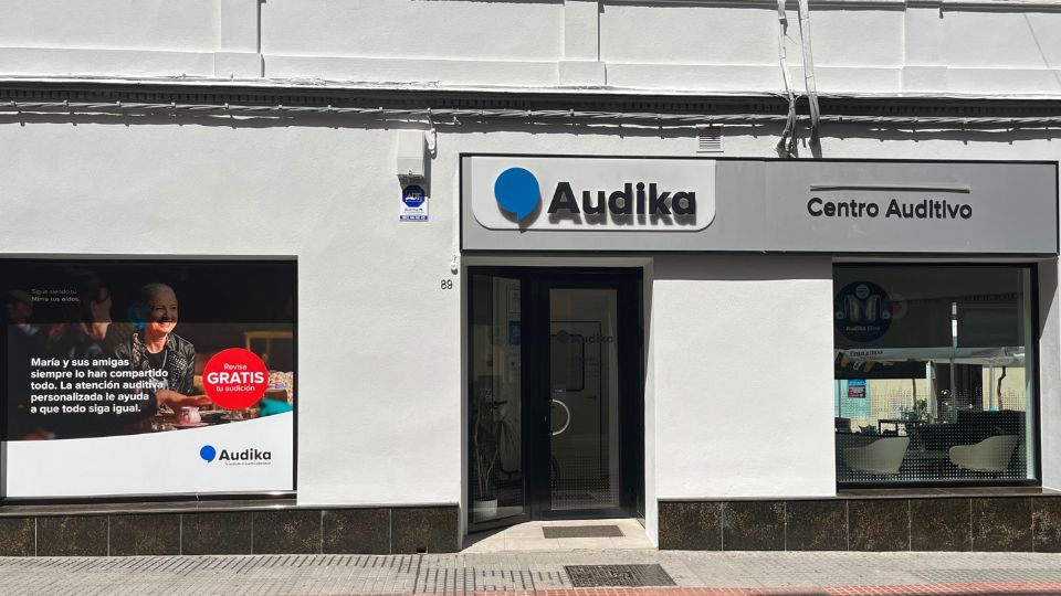 Fachada de un Centro Auditivo Audika con rótulos claros. A la izquierda, un cartel con el texto: ’María y sus amigas siempre lo han compartido todo. La atención auditiva personalizada le ayuda a que todo siga igual. Revisa GRATIS tu audición.’ Inside se observan sillas modernas en un ambiente iluminado.
