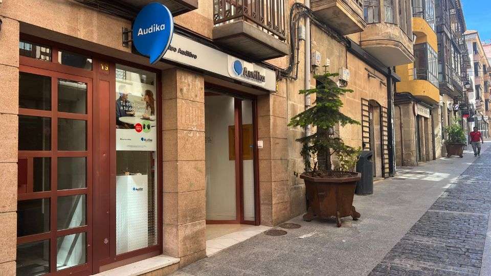 Fachada de una clínica auditiva Audika con puerta y ventana de cristal marrón, ubicada en una calle peatonal con edificios históricos. Un cartel dice: ’Tu oído siempre a tu lado’.