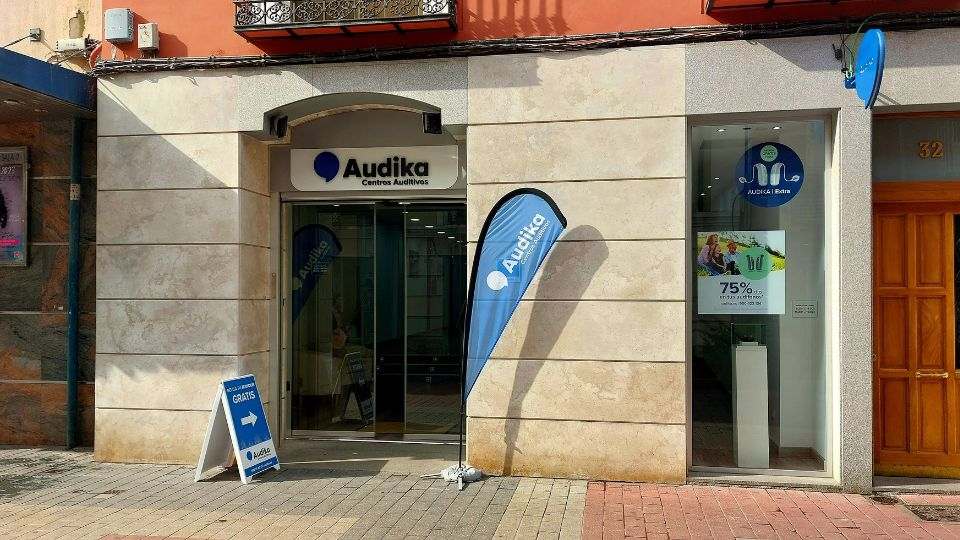 Fachada de una clínica auditiva Audika con entrada de cristal, banderola azul promocional y cartel de prueba auditiva gratuita. En la vitrina, texto: ’75% de descuento en audífonos.’