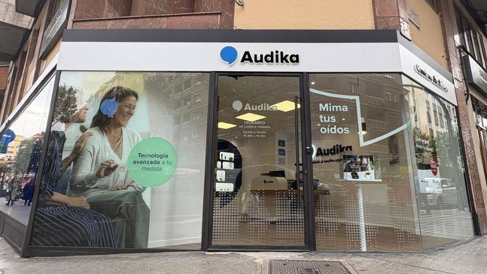 Fachada de una clínica Audika con cristales decorados, destacando textos como ’Tecnología avanzada a tu medida’ y ’Mima tus oídos’. Interior visible con mobiliario para atención en servicios de audífonos.