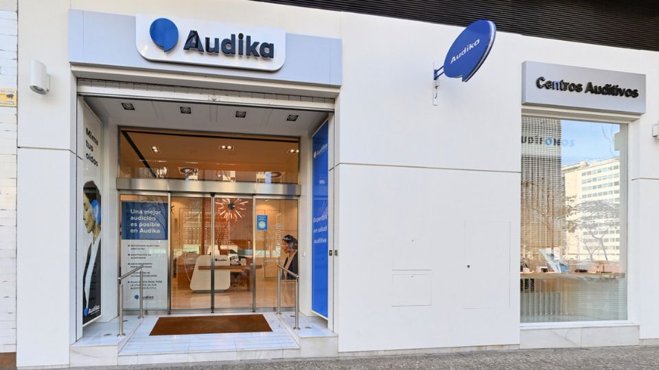 Fachada de una clínica auditiva Audika con puertas de vidrio, iluminación cálida y diseño moderno; incluye texto: ’Una mejor audición es posible en Audika’ y ’Centros Auditivos’.