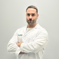 Un profesional de la audición con bata blanca, brazos cruzados y el logo de Audika visible en el pecho, posando contra un fondo gris liso.