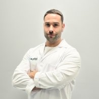 Un profesional de la audición con bata blanca, brazos cruzados y el logo de Audika visible en el pecho, posando contra un fondo gris liso.