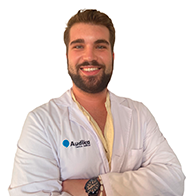 Un audiólogo con bata blanca, brazos cruzados, y el logo de Audika en el pecho; fondo blanco resaltando un entorno profesional de clínica auditiva enfocada en audífonos y cuidado auditivo.