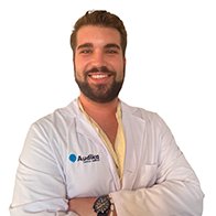 Un audiólogo con bata blanca, brazos cruzados, y el logo de Audika en el pecho; fondo blanco resaltando un entorno profesional de clínica auditiva enfocada en audífonos y cuidado auditivo.