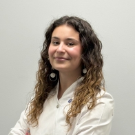 Una profesional de la salud auditiva con bata blanca, brazos cruzados, cabello rizado y castaño, en un entorno neutro de clínica auditiva. No se observa texto ni dispositivos auditivos visibles.