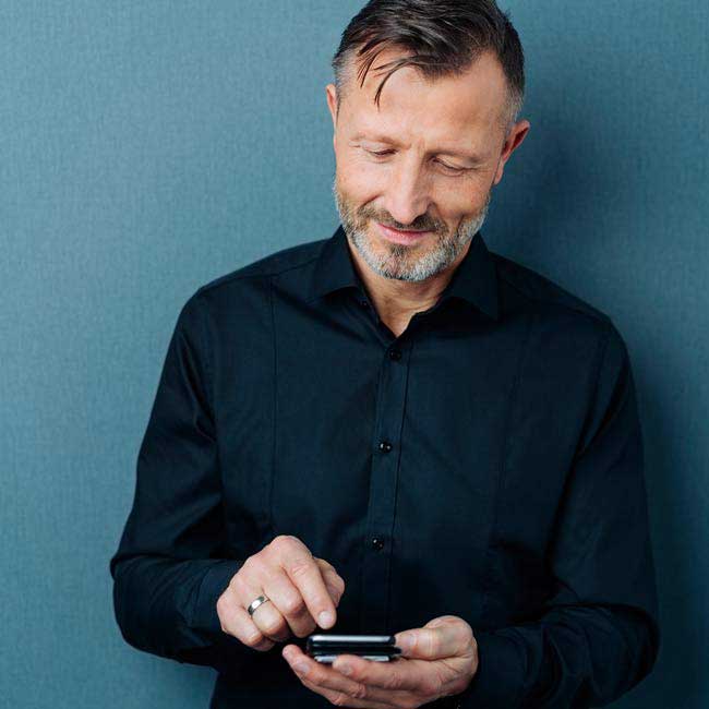 Un hombre con camisa negra interactúa con un teléfono móvil en sus manos, sobre un fondo liso de color azul. No hay texto visible ni elementos relacionados con audífonos.