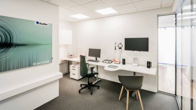 Consultorio moderno de Audika con escritorio, audífonos, pantalla, equipo para pruebas auditivas y sillas, ubicado en una clínica auditiva. En la pared: ’Audika - Para apoyar tu audición’.