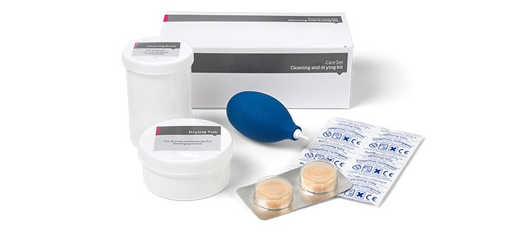 Un kit de limpieza y secado para audífonos compuesto por recipientes blancos etiquetados ’Cleansing Depot’ y ’Drying Tray,’ una bomba de aire azul, dos cápsulas deshumidificadoras, y una caja blanca.