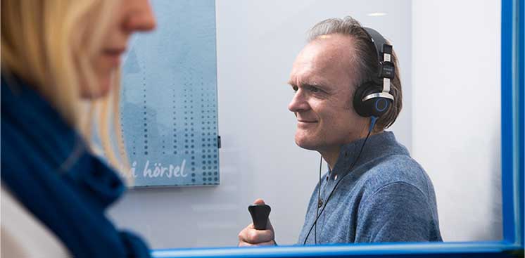Una persona usando audífonos realiza una prueba auditiva en una cabina insonorizada, sosteniendo un pulsador manual. Al fondo, un cartel con texto parcialmente visible: ’hörsel.’ Contexto: clínica auditiva.