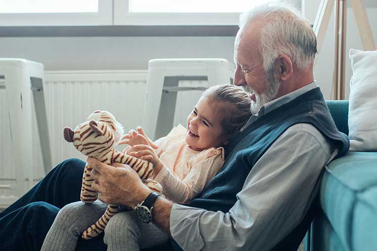 Un hombre mayor sostiene a un niño pequeño mientras ambos manejan un peluche de cebra; están sentados en una sala iluminada con taburetes blancos y un radiator de fondo.