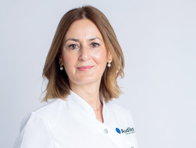 Una profesional de Audika, con uniforme blanco que tiene el logo de la empresa visible. Ella está en un fondo claro, representando un entorno de clínica auditiva especializado en audición y cuidado auditivo.